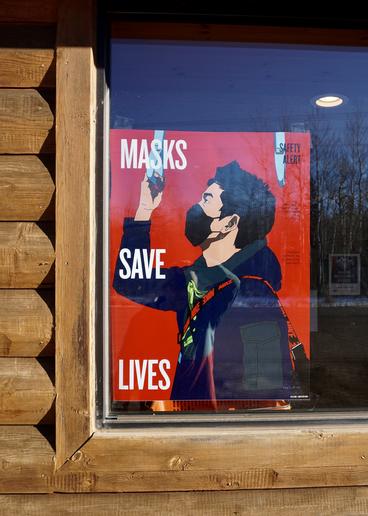 Photo of a sign hanging inside the window of a log building that says masks save lives
